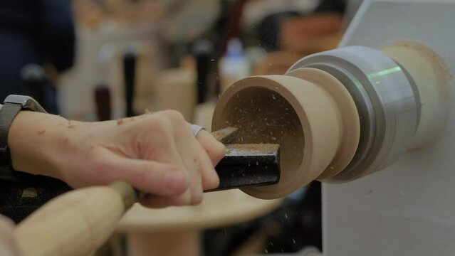 Slow motion: carpenter using chisel for shaping piece of wood on turning lathe machine with many shavings, making wooden mug at workshop - close up. Hobby, carpentry and manufacturing concept