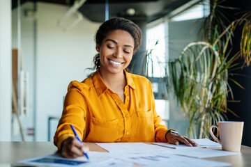 Happy black female company worker working with documents, writing and doing paperwork, sitting at workplace in office - obrazy, fototapety, plakaty