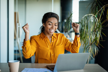 Business success. Overjoyed black woman using laptop and gesturing yes, shaking fists celebrating...