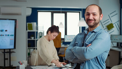Obraz premium Portrait of confident man entering frame posing with arms crossed and smiling in busy startup office. Relaxed employee acting casual looking at camera in marketing department of small company.