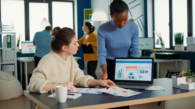 African American Woman Holding Laptop Asking Colleague Looking At Clipboard With Charts For Advice Regarding Financial Data In Busy Office. Two Colleagues Analzying Market Share Brainstorming Ideas.