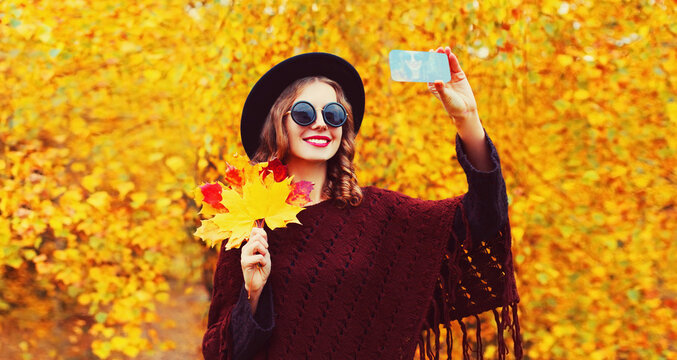 Autumn portrait of stylish happy smiling young woman taking selfie with smartphone and yellow maple leaves wearing round hat, brown knitted poncho in the park