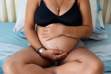 Close up view of a pregnant woman touching and holding her belly while sitting on the bed. Pregnancy concept.