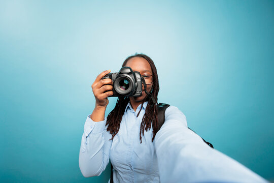 Young adult african american woman pointing DSLR device to camera while taking a photo on blue background. Creative amateur photographer taking pictures with modern camera for social media.