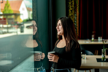 Beautiful girl stands near the window with coffee and smiles
