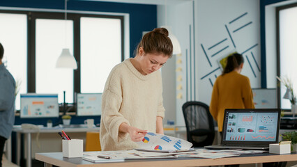 Busy woman in startup office holding clipboard looking at papers with sales charts at desk....