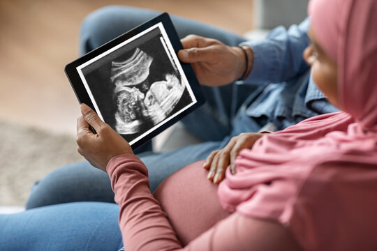 Pregnant black islamic couple holding digital tablet with baby ultrasound image