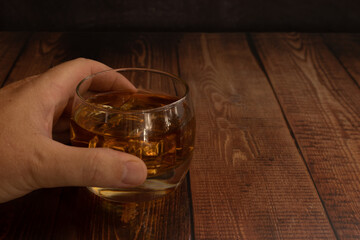 hand of a person holding a glass of whisky rum liquor with ice cubes wooden table with space for text