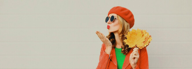 Autumn portrait of beautiful young woman with yellow maple leaves blowing her lips with red lipstick wearing french beret on gray background