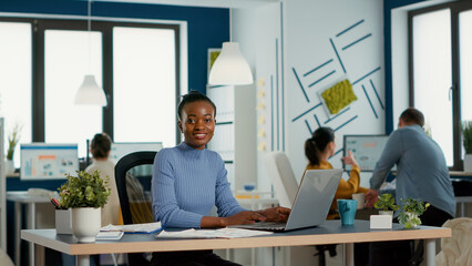 Portrait of smiling african american woman working on profit margin and sale statistics using laptop sitting at desk in busy startup office. Casual business employee happy with job result. - obrazy, fototapety, plakaty