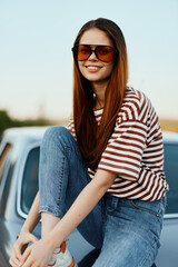 A fashion woman in stylish sunglasses, a striped t-shirt and jeans sits on the trunk of a car and looks at the beautiful nature of autumn. Travel lifestyle