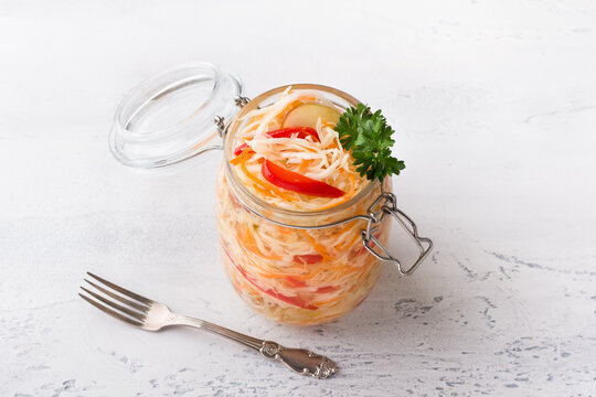 A jar of quick pickled cabbage with carrots, apples and red bell peppers, garnished with parsley on a light blue background. The concept of delicious homemade vegan food