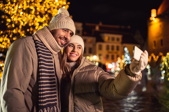 winter holidays and people concept - happy smiling couple taking selfie with smartphone over christmas tree lights in evening city