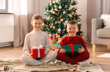 christmas, winter holidays and childhood concept - happy girl and boy in pajamas with gifts sitting on floor at home