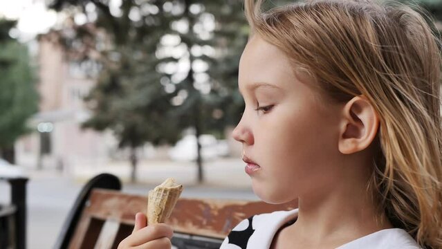 teenager cute caucasoid girl eating ice cream sitting on a bench in the city with her back to the road. The child enjoys ice cream holding an ice cream cone in his hands and biting off dessert