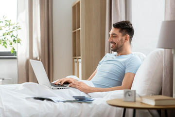 people, technology and remote job concept - man with laptop computer lying in bed at home bedroom