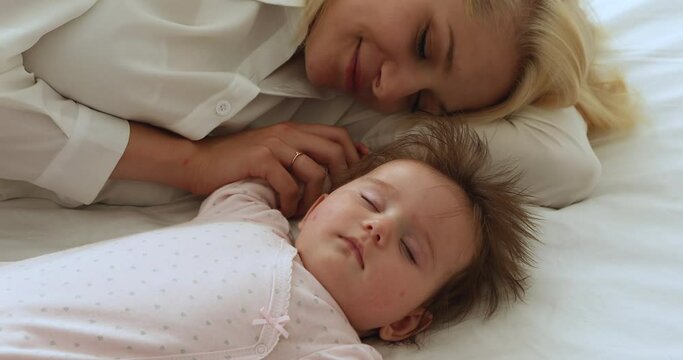 Serene baby and young mother sleeping in bed, close up. Newborn and mom lying down with eyes closed looking carefree and peaceful resting together at home. Sweet dreams, maternity, co-sleeping concept