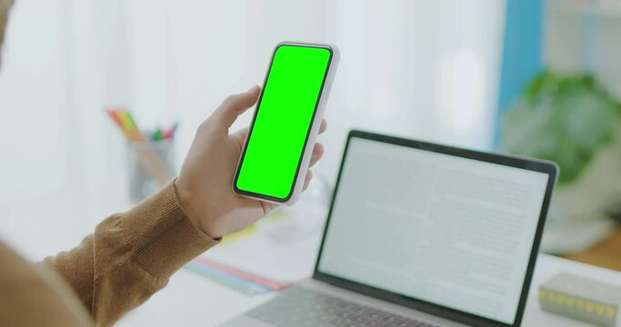 Over The Shoulder Locked Shot of Man Hand Holding and Clicking on Smartphone with Green Screen. Tracking Points to Insert You Own Content. Modern Desk With Laptop In The Background. 