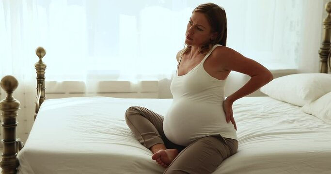 Pregnant woman sits on bed, massages her aching lower back. Unhealthy expectant mother suffers from severe backache due to big belly or overweight during hard pregnancy, having health problem concept