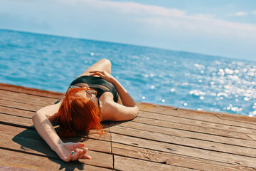 Woman in a swimsuit is lying on a wooden pier and sunbathing. Red-haired girl is enjoying a vacation at the sea. Summer trip. Copy space for travel agency advertising.