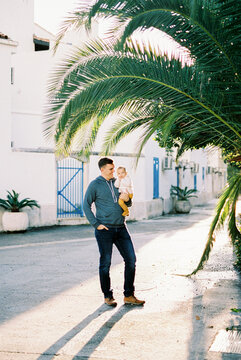 Dad With A Baby In His Arms Stands Near Huge Palm Leaves In The Courtyard Of The House