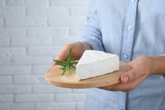 Woman Holding Serving Board With Delicious Brie Cheese Near White Brick Wall, Closeup