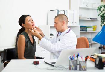 Focused therapist sitting at table in medical office, examining tonsils of Asian woman - obrazy, fototapety, plakaty