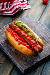 Hot dog American with cucumbers and tomatoes and ketchup on board on blue wooden table vertical