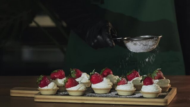 Chef sprinkling sugar powder on tasty mini cake with cream and strawberries, slow motion. Powdered sugar being sieved on strawberry tart. Dessert dusted with castor sugar