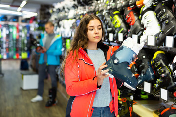 Fototapeta na wymiar Young customer woman in ski equipment is choosing boots for skiing in store
