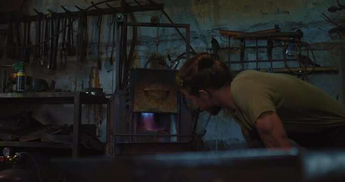 young blacksmith is waiting heating of furnace in smithy, craft workshop, 4K, Prores