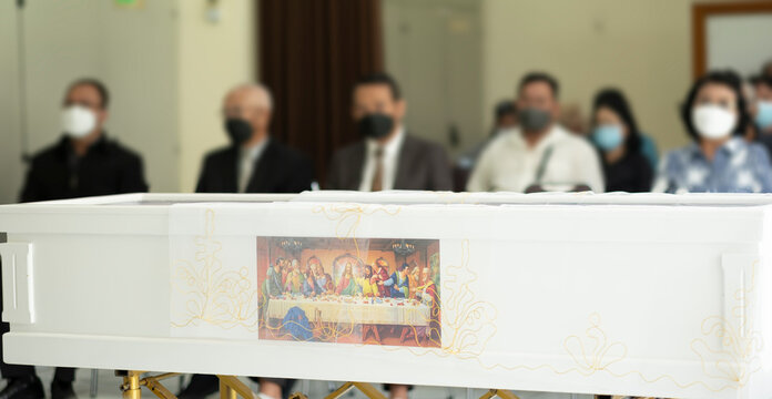 Funeral ceremony during Covid-19 pandemic. Grieving people with mask are sitting in front of a white coffin as blur background. A white casket with picture of the last supper covered by white vitrage.
