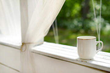 White mug with tea or coffee on a white wooden table on summer terrace in green garden, blurred green background, copy space