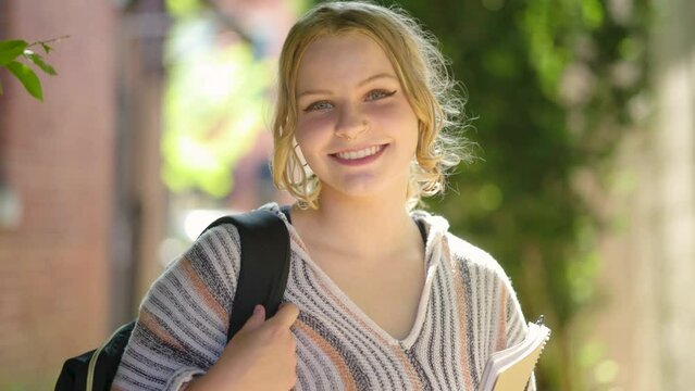 Girl college student holding a backpack and notepad smiles at the camera. Slow motion.