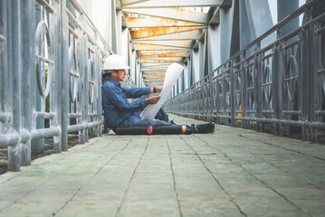 Asian male engineer sitting on the bridge and looking at blueprint for planning to improve the structure, to people and engineering concept.