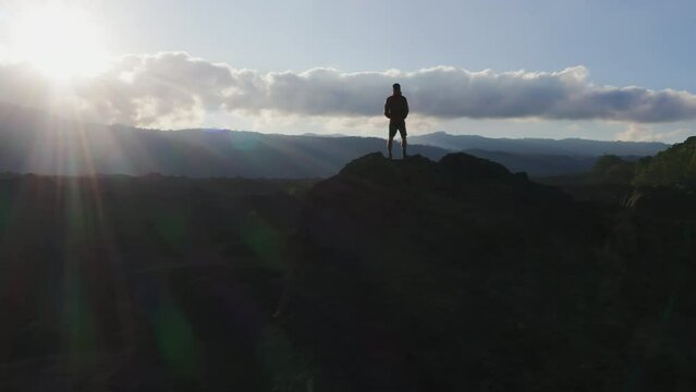 A man standing on a mountain peak in rays of evening sun. Aerial drone footage of a male traveler on top of mount in highlands at sunset. Achieving goals metaphor. Hero on a rock. Hiking trip moments.