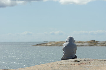 A lonely boy in a grey hoodie sits on a rock looking out over the sea