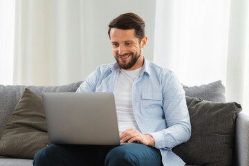 Man working or studying online remotely using laptop. Positive attractive caucasian male sitting on sofa at home using computer, smiling