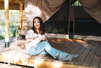 Brunette woman in nordic sweater drinking tea and relaxing in glamping in nature