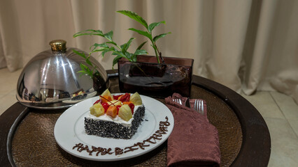 On the coffee table, on a white plate, there is a cake decorated with cream and fruit. There is a congratulatory inscription on the rim. Next to it is a metal lid, cutlery, a plant in a vase.