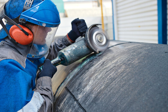 Linear fitter or pipeliner removes plastic insulation from large-diameter pipe with angle grinder. Young worker in overalls and protective mask works outside and processes metal pipe.