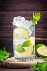 Homemade sweet and sour cold lemonade or infused water made of lime and lemon slices, mint leaves, soda and ice cubes served in glass on dark brown wooden table used as organic refreshing drink