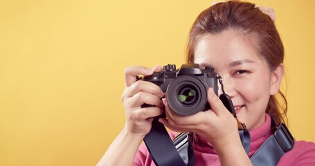 Young Asian woman playing in pink clothing using a digital camera against an isolated yellow background. Travel concept