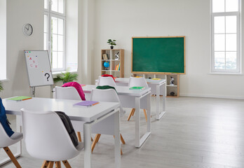 Bright interior of empty classroom without students in modern elementary school. School classroom with blackboard, white desks and chairs with colored backpacks. Educational concept.