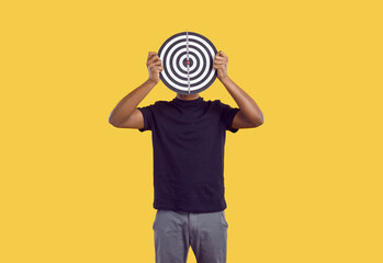 Unknown dark-skinned man in casual clothes hides his face behind darts board on yellow background....