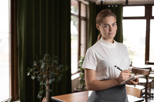 Happy female waiter in apron writing order in restaurant
