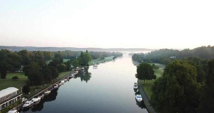 Beautiful drone shot of Henley on Thames at sunrise, flying along the river to the East with a light mist over the river and surrounding hills and fields.