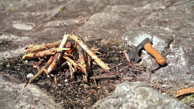 Small bonfire starting to burn in outdoor rocky fire pit with small hiking axe laying by its side - Sunny day outdoor static clip from nature - Flames moving in slow motion
