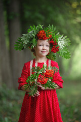 a blond preschool girl in a wreath of rowan and with toys walks in the park on a summer morning