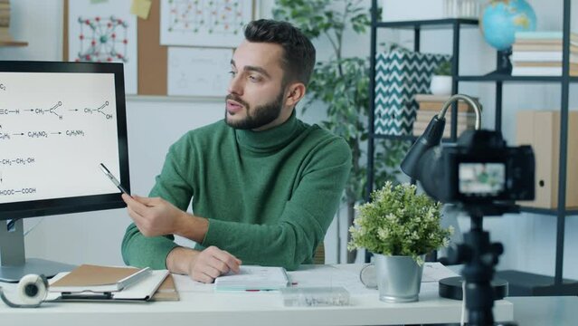 Young Scientist Recording Video About Chemistry Using Modern Camera In Home Office. Man Pointing At Computer Screen And Speaking Giving Lecture.